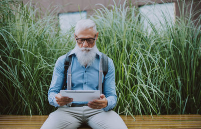 Senior hipster holding digital tablet sitting against plants