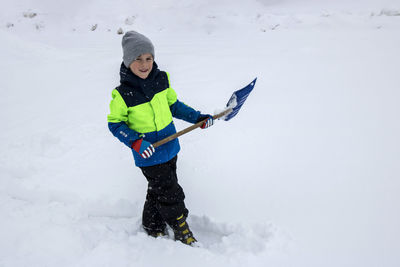 Full length of cute boy holding shovel while standing on snowy land during winter