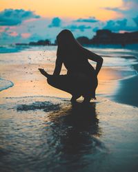 Silhouette woman crouching at beach against cloudy sky during sunset