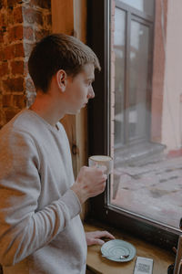 A teenager in a white hoodie looks thoughtfully out the window, stands with a cup of coffee 
