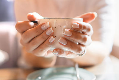 Closeup of female hands with french manicure holding cozy ceramic white mug of tea or coffee