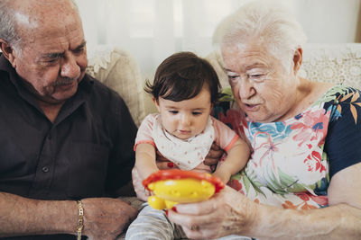 Great-grandparents sitting with baby girl on the couch at home looking at toy