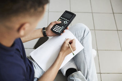 Young man using calculator and writing on paper while sitting with crossed legs at laundromat