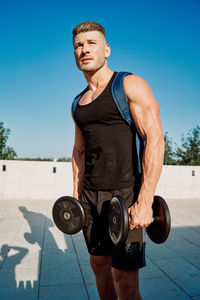 Portrait of man exercising with dumbbells against sky