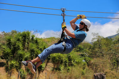 Woman wearing helmet and gloves, zip lining in tropical setting.