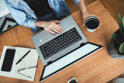 High angle view of woman sitting at desk and working on laptop while holding coffee mug