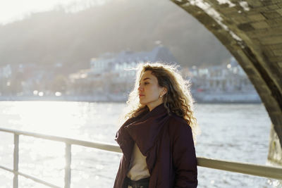 Portrait of young woman standing under the bridge against river 