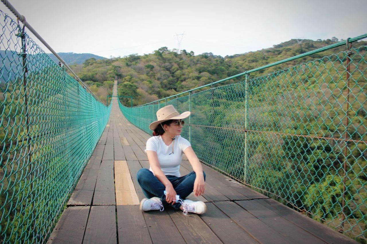 one person, full length, hat, nature, adult, fence, clothing, leisure activity, sitting, day, lifestyles, casual clothing, sun hat, women, green, sky, relaxation, outdoors, footwear, sports, plant, young adult, person, summer, environment, front view, fashion, trip, vacation, chainlink fence, copy space, looking, landscape, holiday, emotion, architecture, water