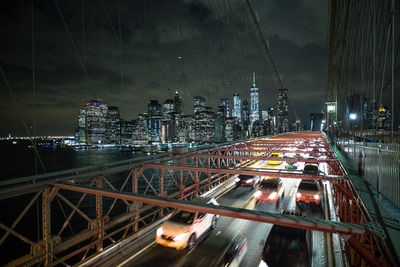 Light trails on bridge by buildings against sky at night