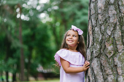 A beautiful girl in a pink dress stands near a tree in the forest in summer, smiling and dreaming