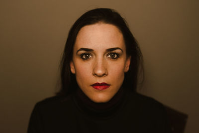 Serious young brunette looking at camera thoughtfully while standing against brown wall