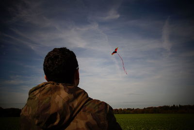 Rear view of army soldier looking at kite on grassy field against sky