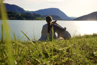 Woman sitting on grass by lake against mountains