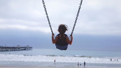 Rear view of girl playing on swing at beach against sky