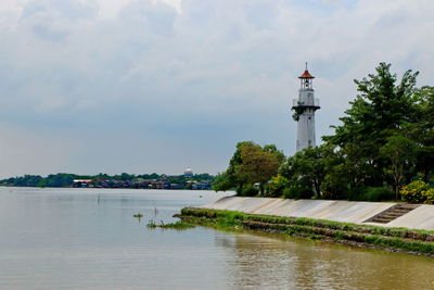 Lighthouse by river and buildings against sky