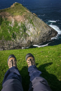 Low section of man sitting on grass field at cliff against sea