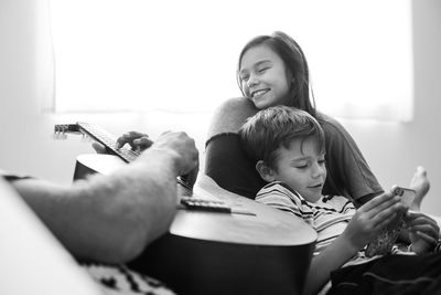 Midsection of father playing guitar while sitting with children against window at home