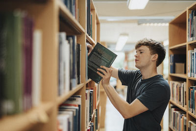 Side view of male student removing book from bookshelf in library at university