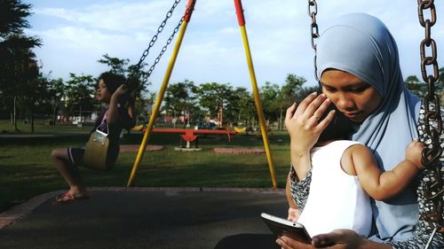 Woman sitting on swing with daughter while using mobile phone at park
