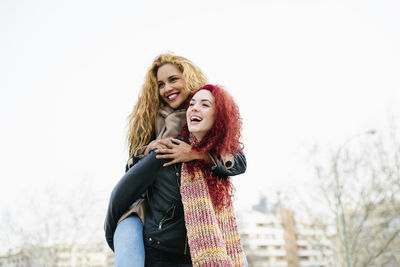 Happy young woman piggybacking girlfriend against clear sky