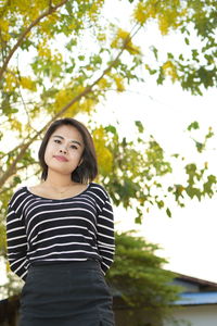 Low angle portrait of beautiful young woman standing against trees in park