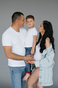 Pregnant couple, a young family with a young son, stand on a gray background, laughing and waiting