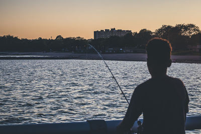 Rear view of silhouette man standing by sea against sky during sunset