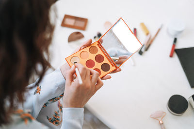 Young brunette woman in blue pajamas applies makeup with face sculpting palette in hands at home