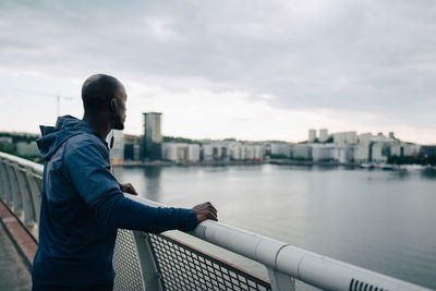 Side view of sportsman looking at city while standing on footbridge over sea against sky