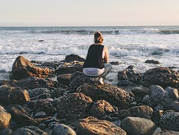 Full length rear view of woman crouching on rocky sea shore against sky during sunset