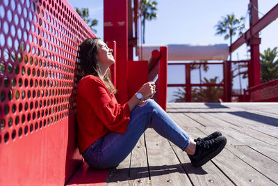 Teenage girl using mobile phone while wearing red top in city against railing during sunny day