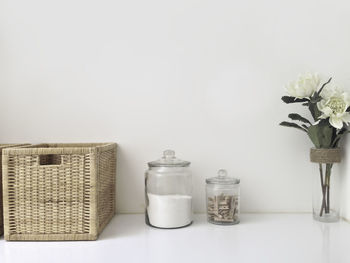 Close-up of basket by jars and vase on table against white background