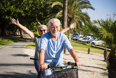 Portrait of senior man with woman riding bicycle on road at park during sunny day
