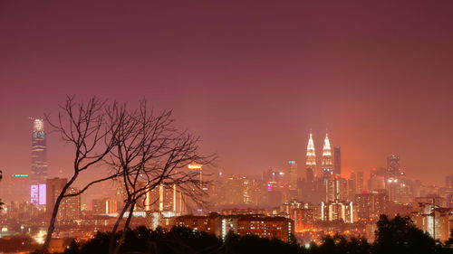 Bare tree and illuminated buildings in city against clear sky at night