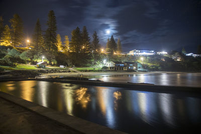 Illuminated city by river against sky at night
