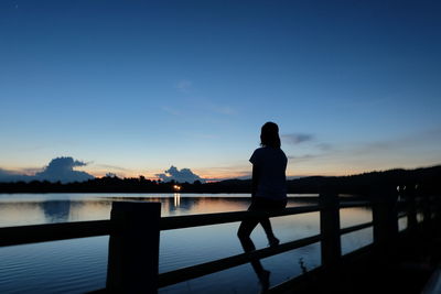 Silhouette woman sitting on jetty against lake at sunset