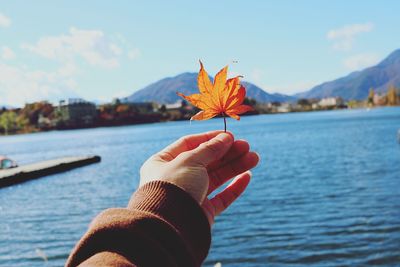 Close-up of hand holding maple leaf by lake during autumn