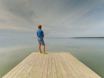 Small blond hair boy is fishing on mole. kid in blue shirt, grey striped shorts and green flip-flops