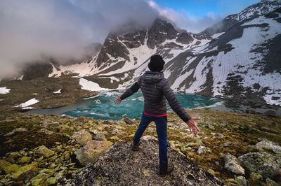 A young man in a downy sweater and panama with his arms outstretched stands in front of a mountain