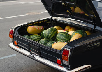 High angle of ripe melons and watermelons for sale placed in opened trunk of vintage black vehicle parked on asphalt road on city street