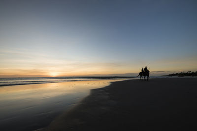 Silhouette men riding horses at beach against sky during sunset