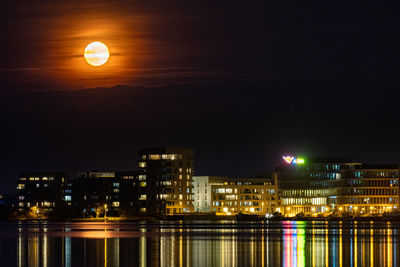 Illuminated buildings by river against sky at night