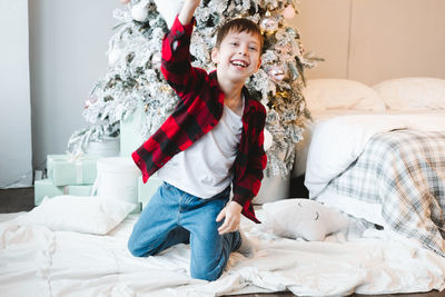 A boy in a red checkered shirt sits by the christmas tree with gifts on the floor 