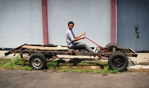 Side view full length of man looking away while sitting on cart against wall