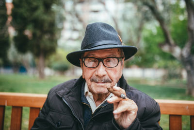 Happy senior male with mustache wearing hat smoking cigar and looking at camera while sitting on bench in park with green trees on blurred background
