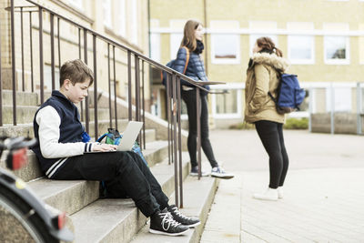 Full length of boy using laptop on steps with schoolgirls in background outside school building