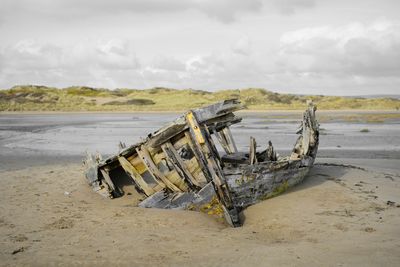 Damaged boat on beach against cloudy sky