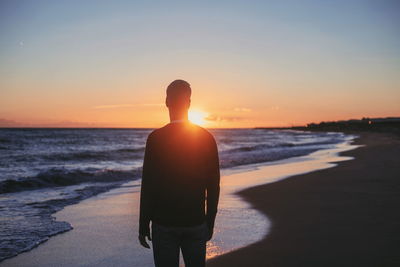 Rear view of man standing at beach against sky during sunset