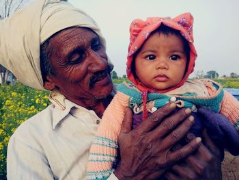 Close-up of mature man carrying granddaughter while standing against sky at farm