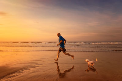 Mature man with dog running at beach against sky during sunset